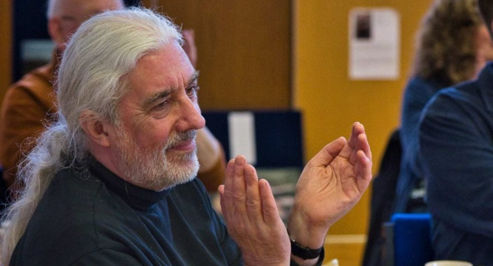 A man with long white hair and beard, clapping his hands.