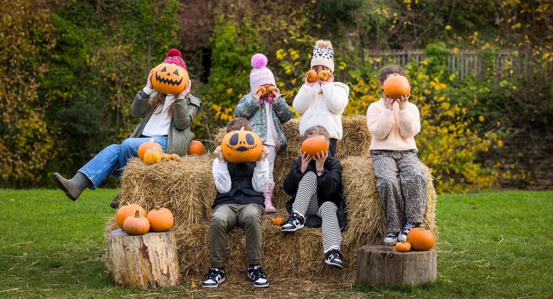 6 children sitting on bales of hale, holding pumpkins in front of their faces.