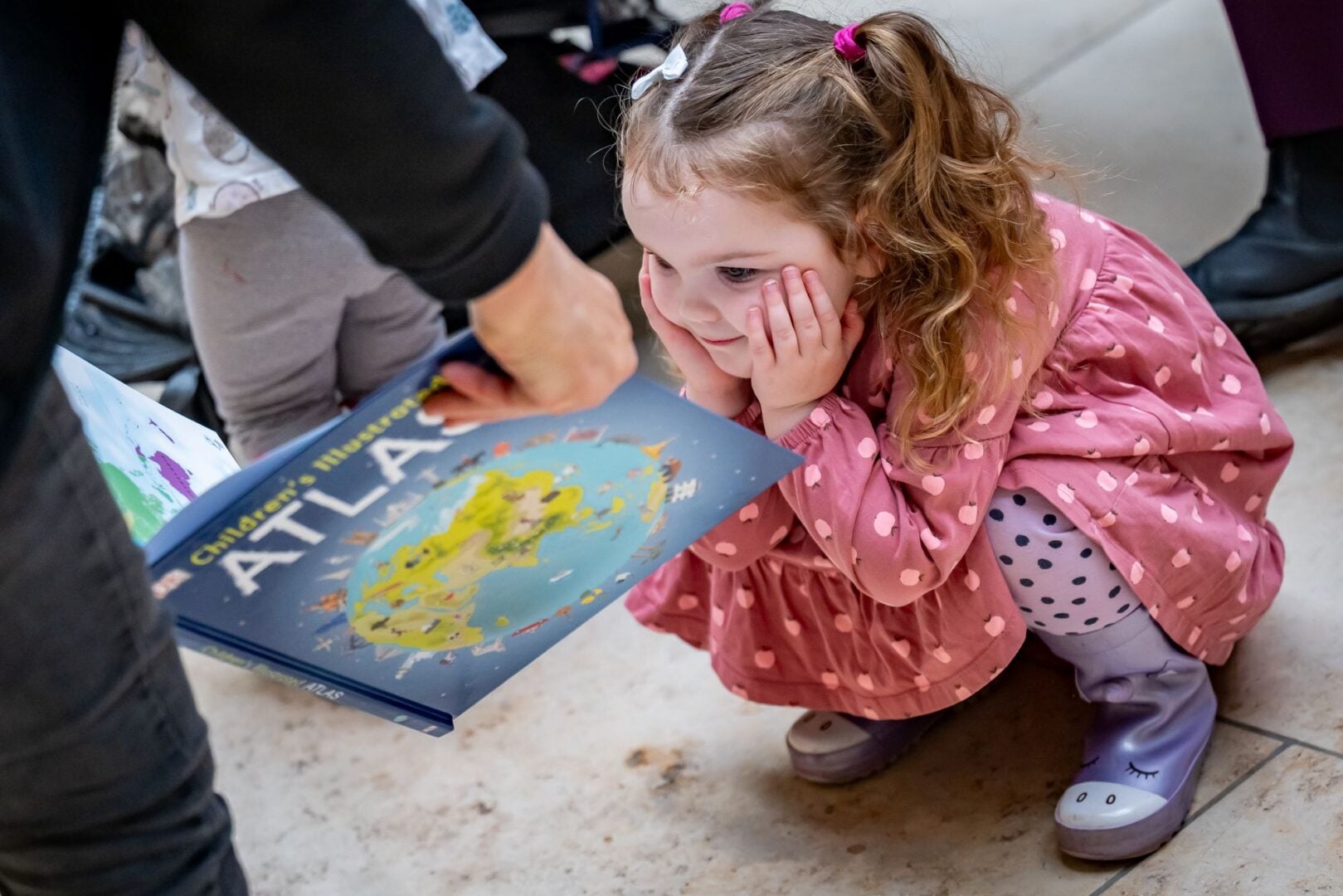 A young girl crouching down to look at an open book that someone is showing her.