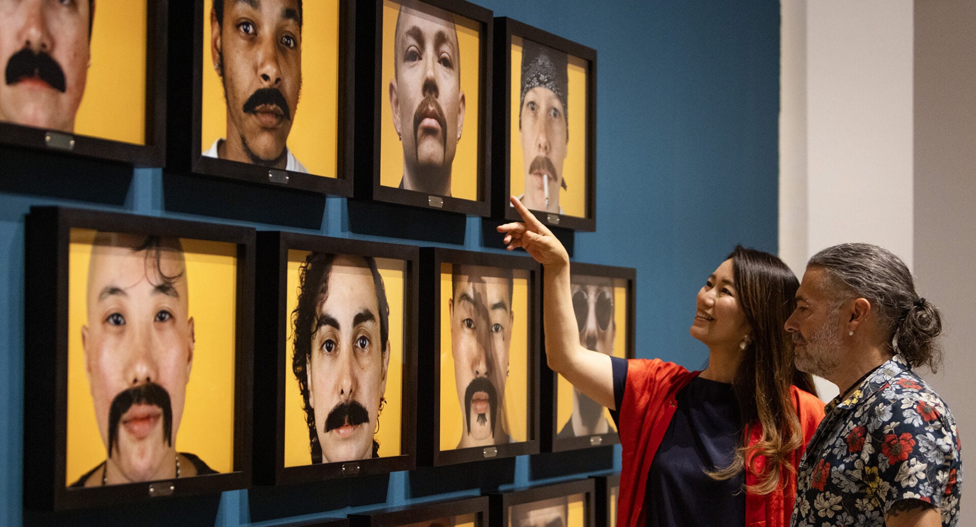 A man and woman looking at a wall on which there are a number of photography portraits.