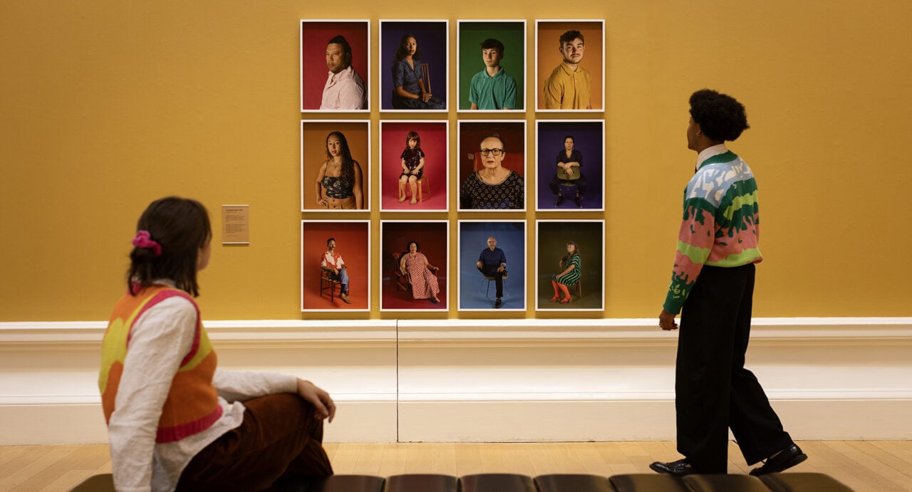 A couple looking at wall, on which there are a collage of portraits.