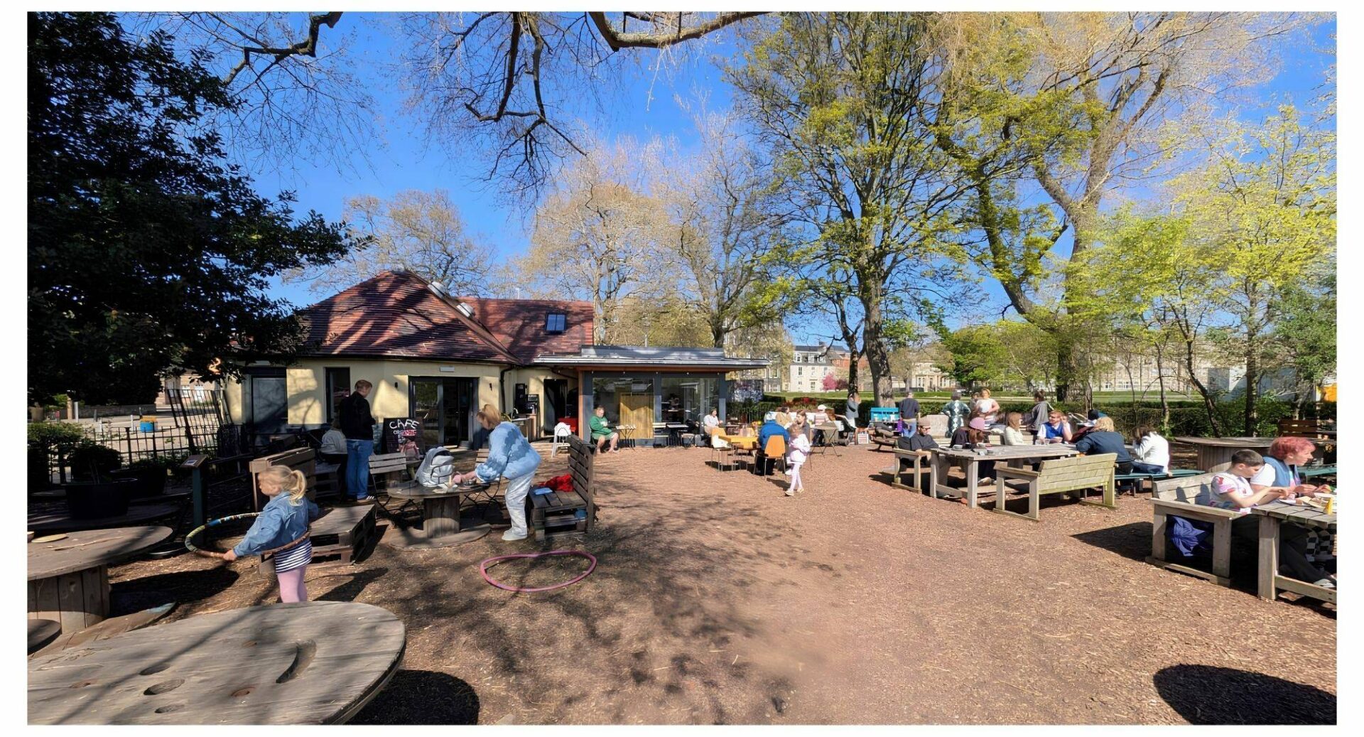 A view of the outside of Populus cafe building, a wide view of the back of the building and large trees. There are groups of people seated inside and outside enjoying coffee and food on a sunny day.,© Earth in Common