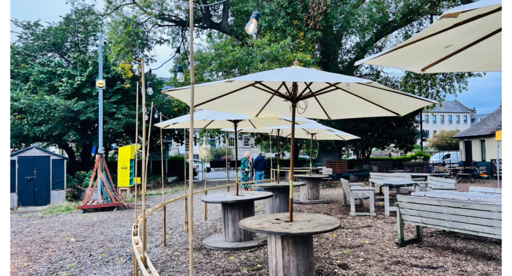 A view of Populus cafe outdoor seating area. long benches and tables, cable reel tables with white shade umbrellas, woodchip on the ground,© Earth in Common