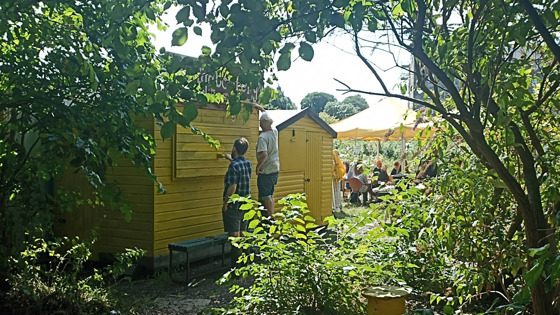 View of the yellow sheds through some trees on Leith Community Croft. Two volunteers are standing in front of the sheds giving them a fresh coat of paint, while people gather under a yellow roffed gazebo in the background,© Earth in Common