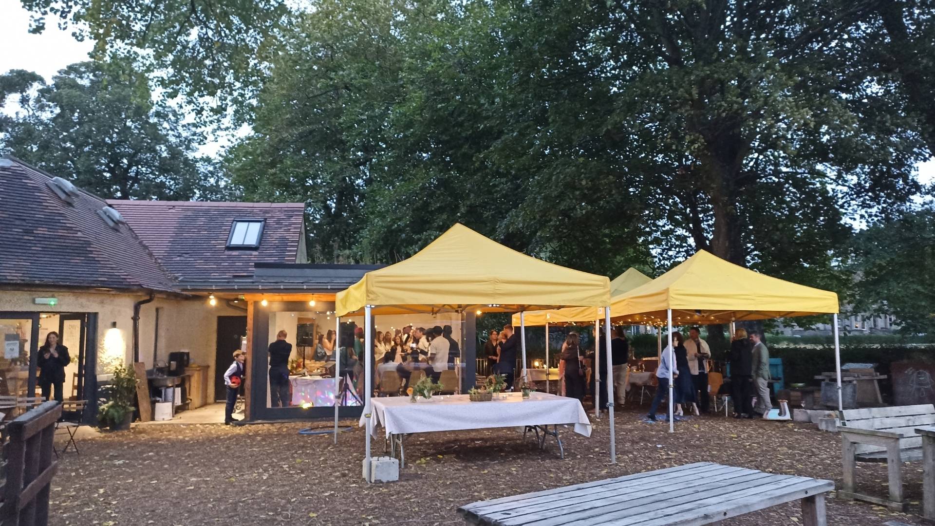 A view of the outside of Populus cafe building, a wide view of the back of the building and large trees. Centrally is a large group of people gathered inside and outside celebrating a wedding.,© Earth in Common