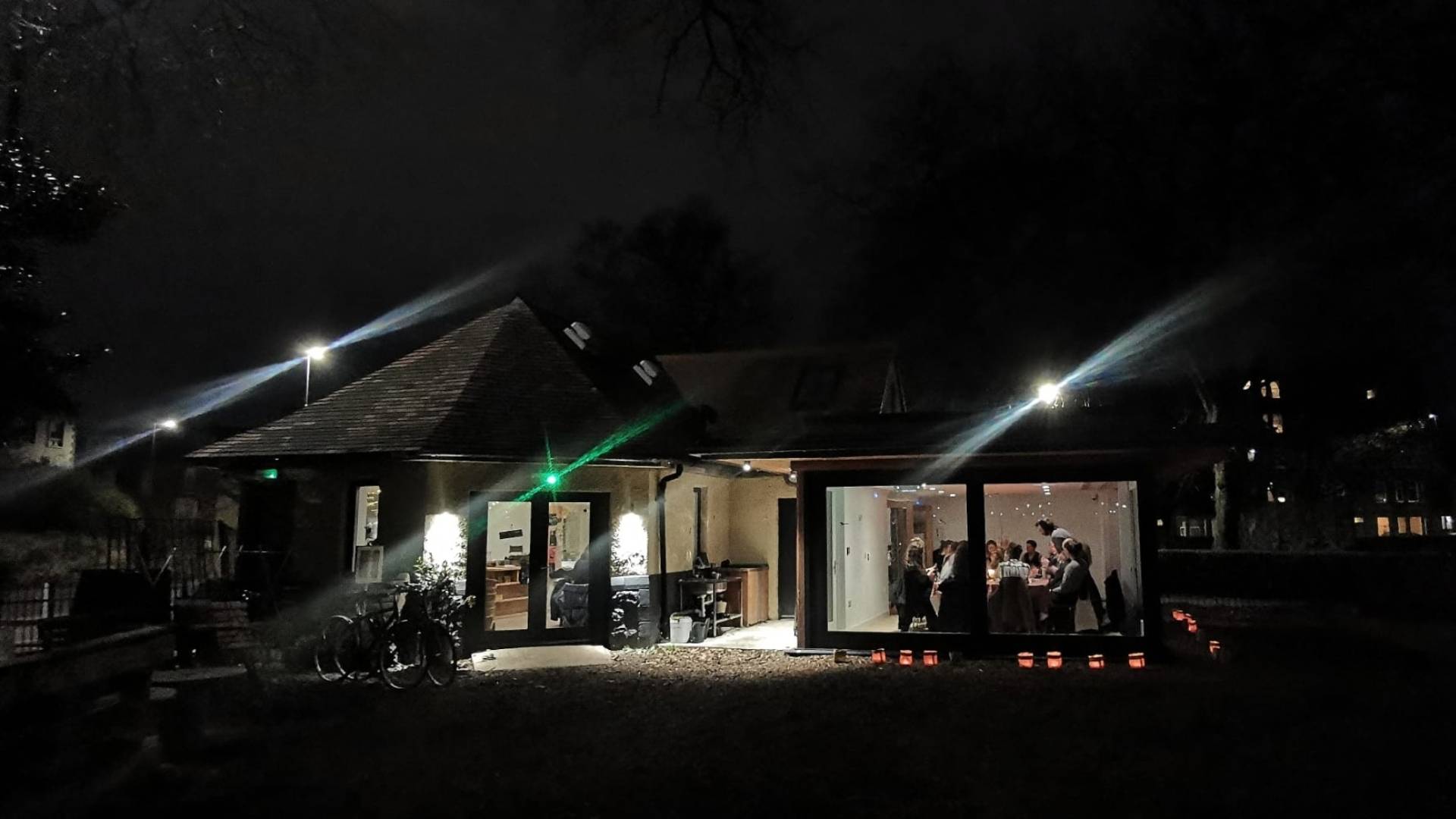 A view of the outside of Populus cafe building, a wide view of the back of the building in the dark. The light is on and a group of people gathered inside around a large table.,© Earth in Common