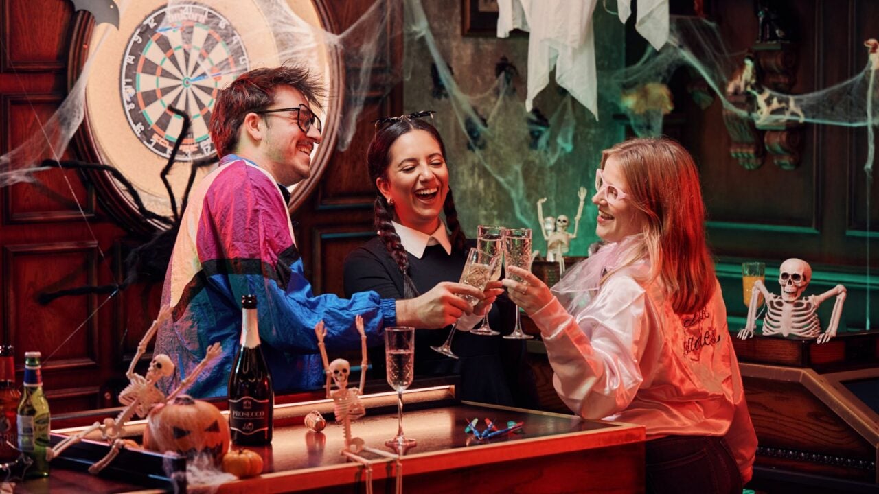 3 people gathered round a bar table, with a dart board in the background.