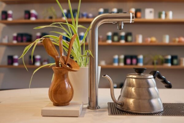 Pekoe Tea counter with a plant, some wooden utensils and a tea pot and tap. In background of image there are shelves.