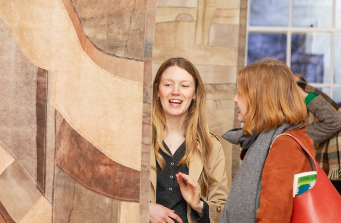 A photograph of two people in conversation. One is facing the camera, the other stands in side profile. The person facing the camera is gesturing with their hands and appears to be in the middle of speaking. They are stood between two long textile sheets that hang from somewhere above them. There is a window in the background. ,© Earth Matters preview, Inverleith House, Royal Botanic Garden Edinburgh, 2026. Photo: Louise King.