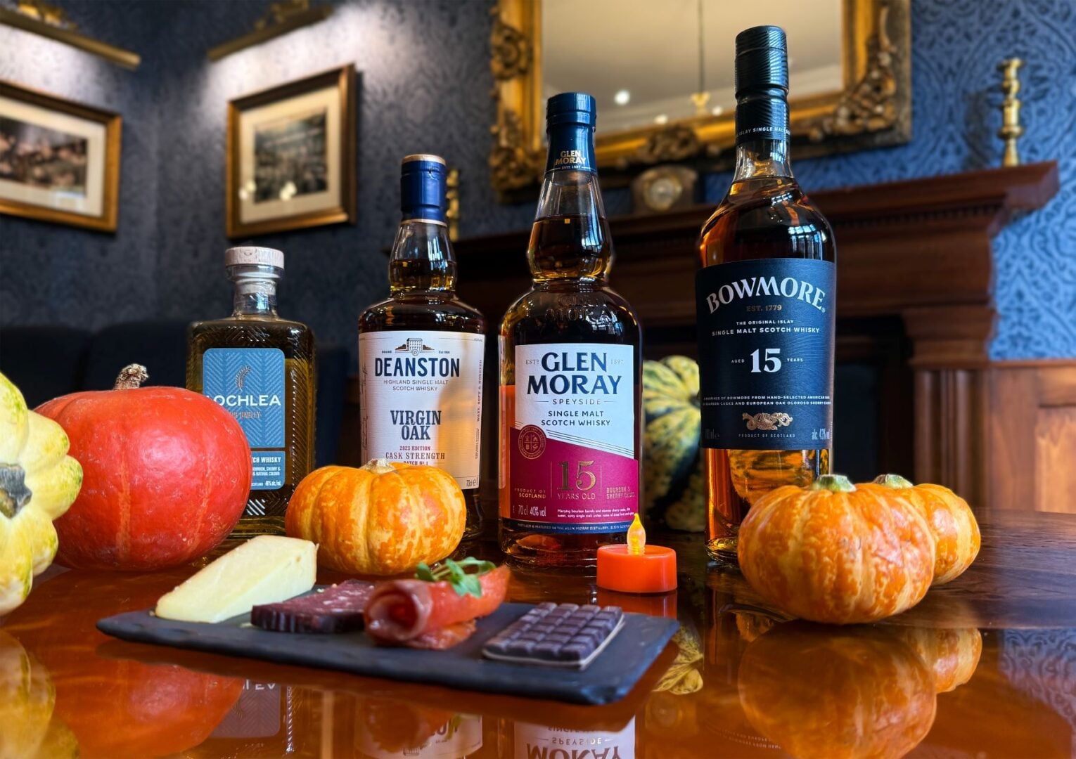 4 bottles of whisky, sitting behind a plate filled with cheese and chocolate, and decorated with small orange pumpkins.