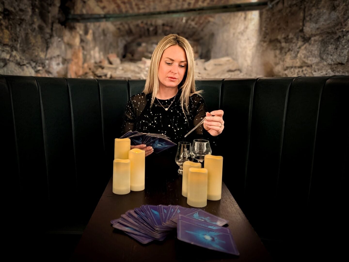 A woman sitting a table. In front of her are 2 whisky glasses, 6 candles and a deck of cards.