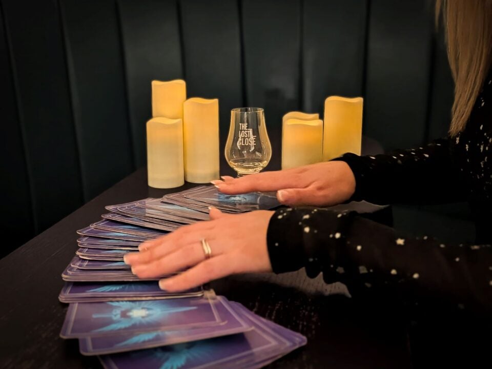 Close-up of a person's hands, fanning out a deck of cards on a table, beside a whisky glass and 6 lit candles.