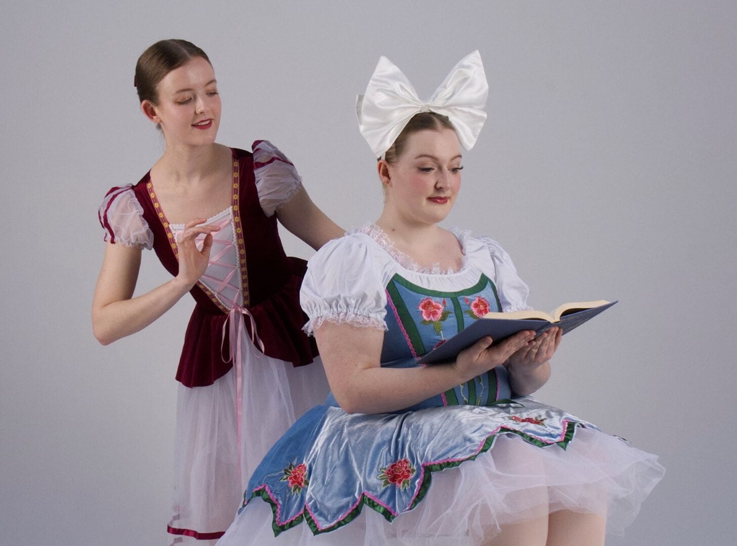 2 young girl, one seated and reading a book, the other standing behind.