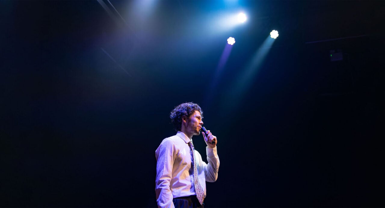 A man holding a microphone, standing on a stage, under a light.