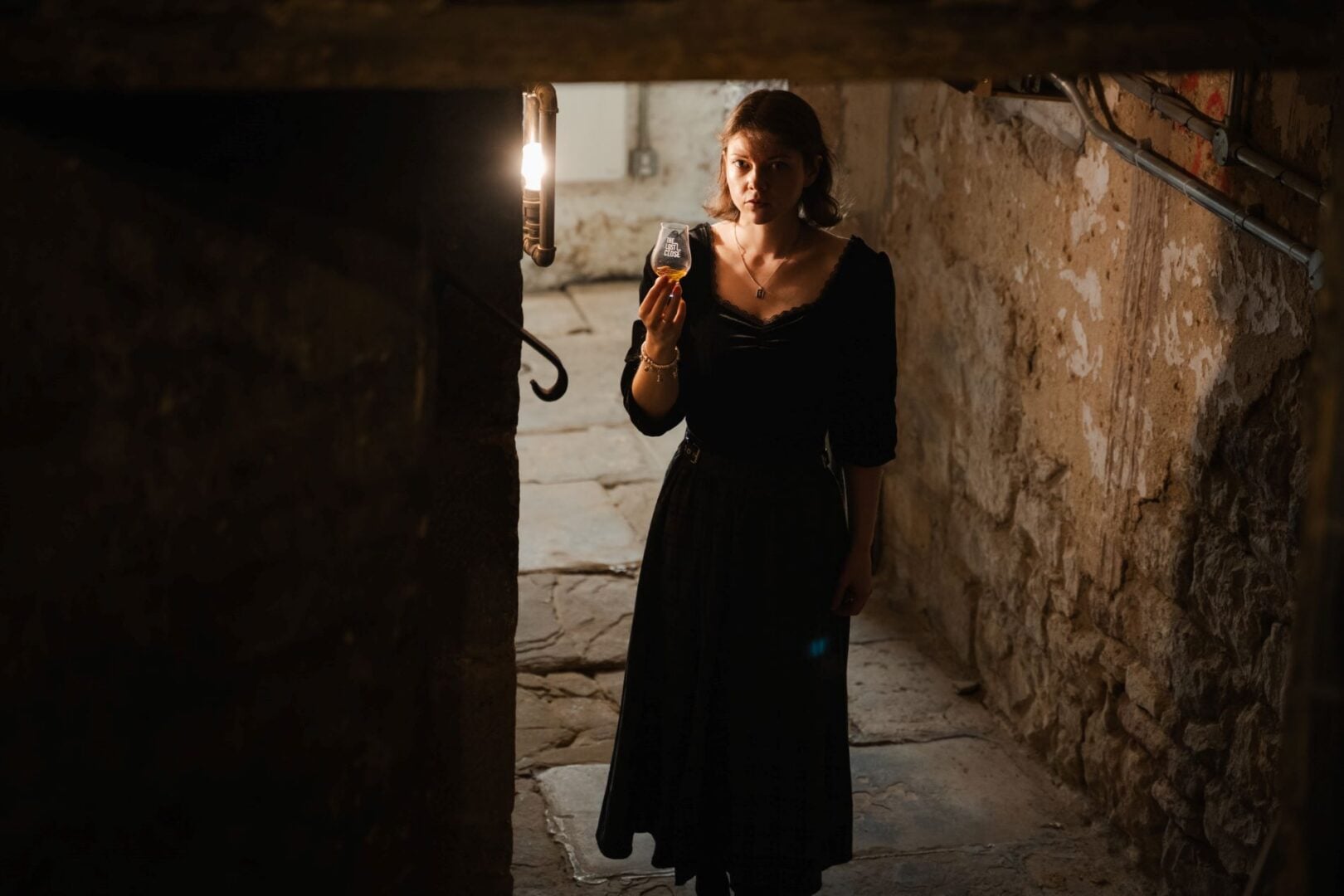 A woman standing in a dimly lit underground space. In her hand is a glass of whisky.