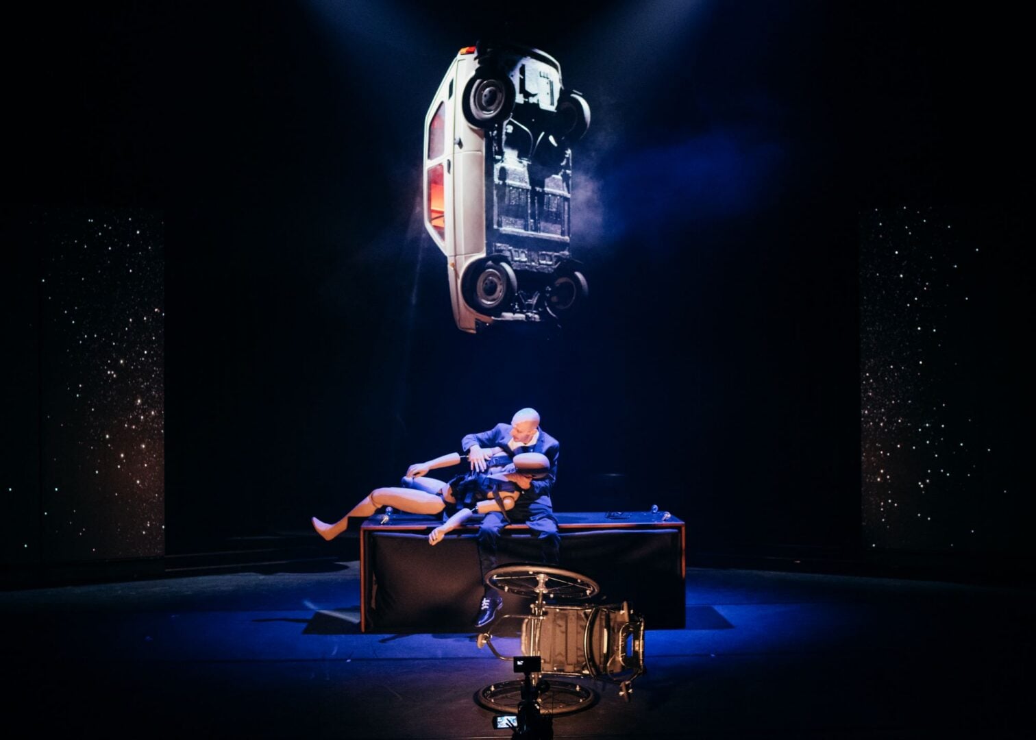 A car hanging in mid air, with 2 people and a wheelchair below it.